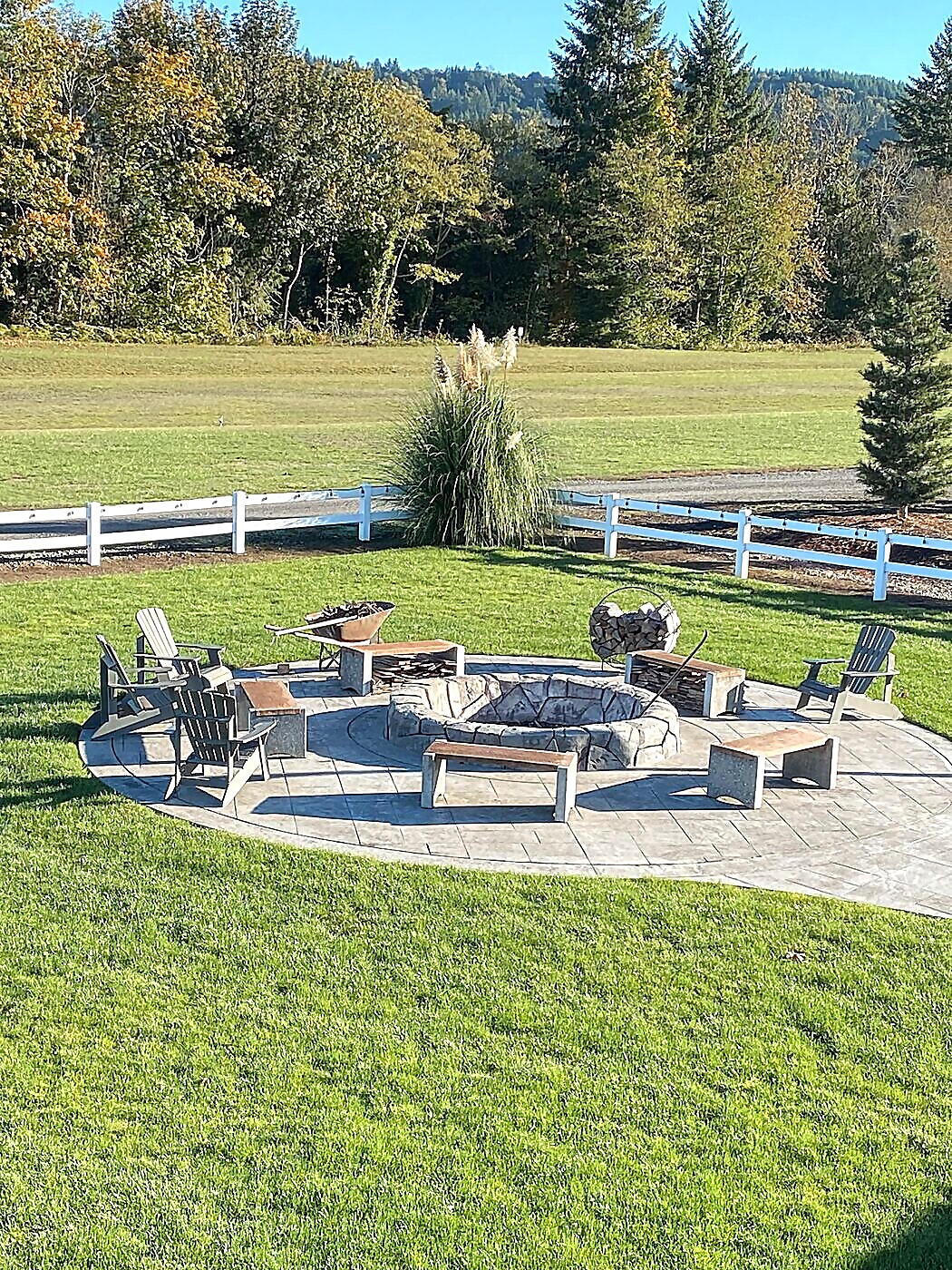 Stone fire pit area with Adirondack chairs and benches at Sandy River Airport, pampas grass and white fence